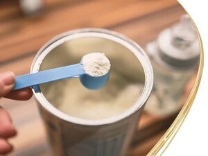 Valokuva kädestä, jossa pidellään sinistä kauhallista valkoista jauhetta avoimen äidinmaidonkorvikkeen päällä